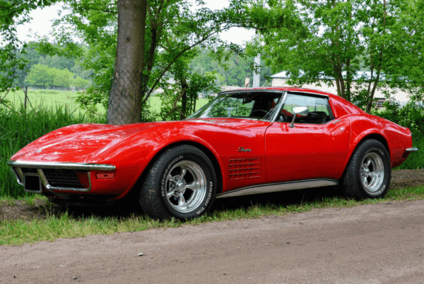 Antique red Chevrolet Corvette Stingray parked on a rural road in Newfoundland, surrounded by green trees and countryside.